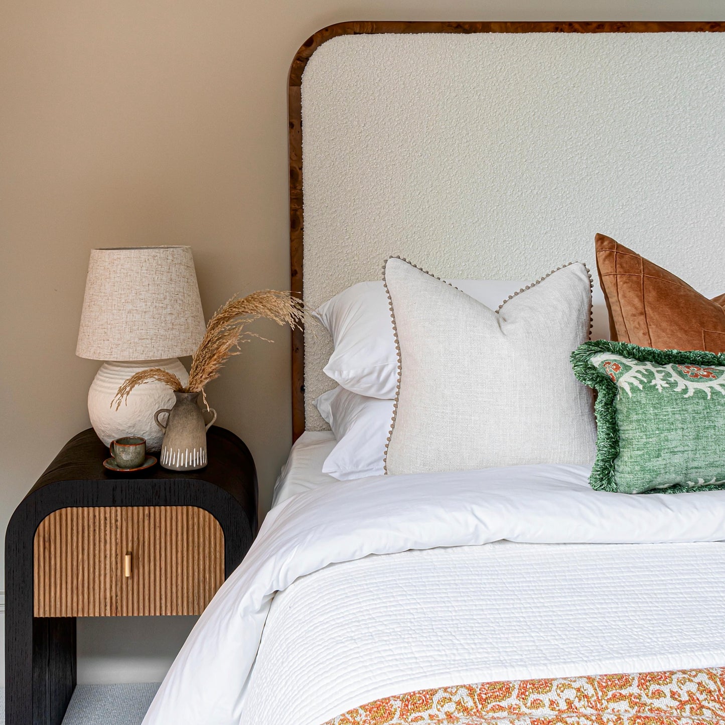 A Thornhill burl wood headboard with white boucle upholstery, positioned behind a bed with white and brown bedding, next to a nightstand with a lamp and decorative objects.